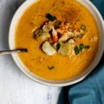a bowl of creamy orange soup with croutons, crawfish tails and green onions