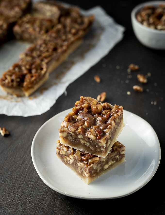 pecan pie bars on a plate with tray of bars and bowl of pecans in the background
