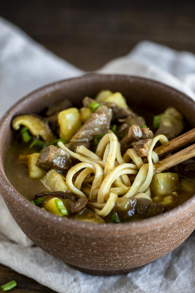 chopsticks in a bowl of noodles with beef and potato in a brown gravy