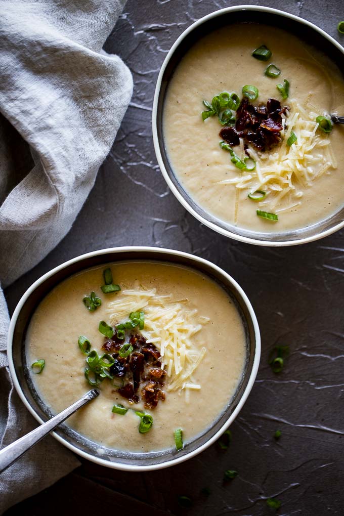 pureed cauliflower soup in bowls with spoons, bacon, green onions and shredded cheese
