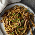 a plate of stir fried noodles with chicken and bacon pieces and chopsticks on the side