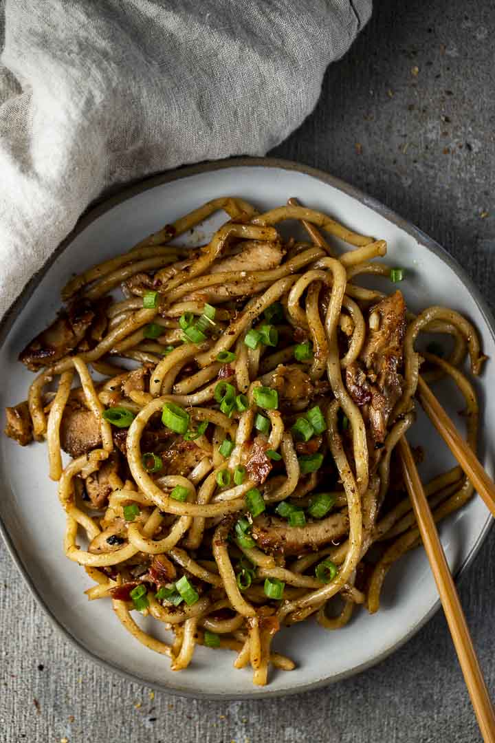 a plate of stir fried noodles with chicken and bacon pieces and chopsticks on the side