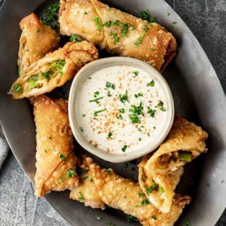 a plate of egg rolls with white dipping sauce garnished with red pepper seasoning and parsley