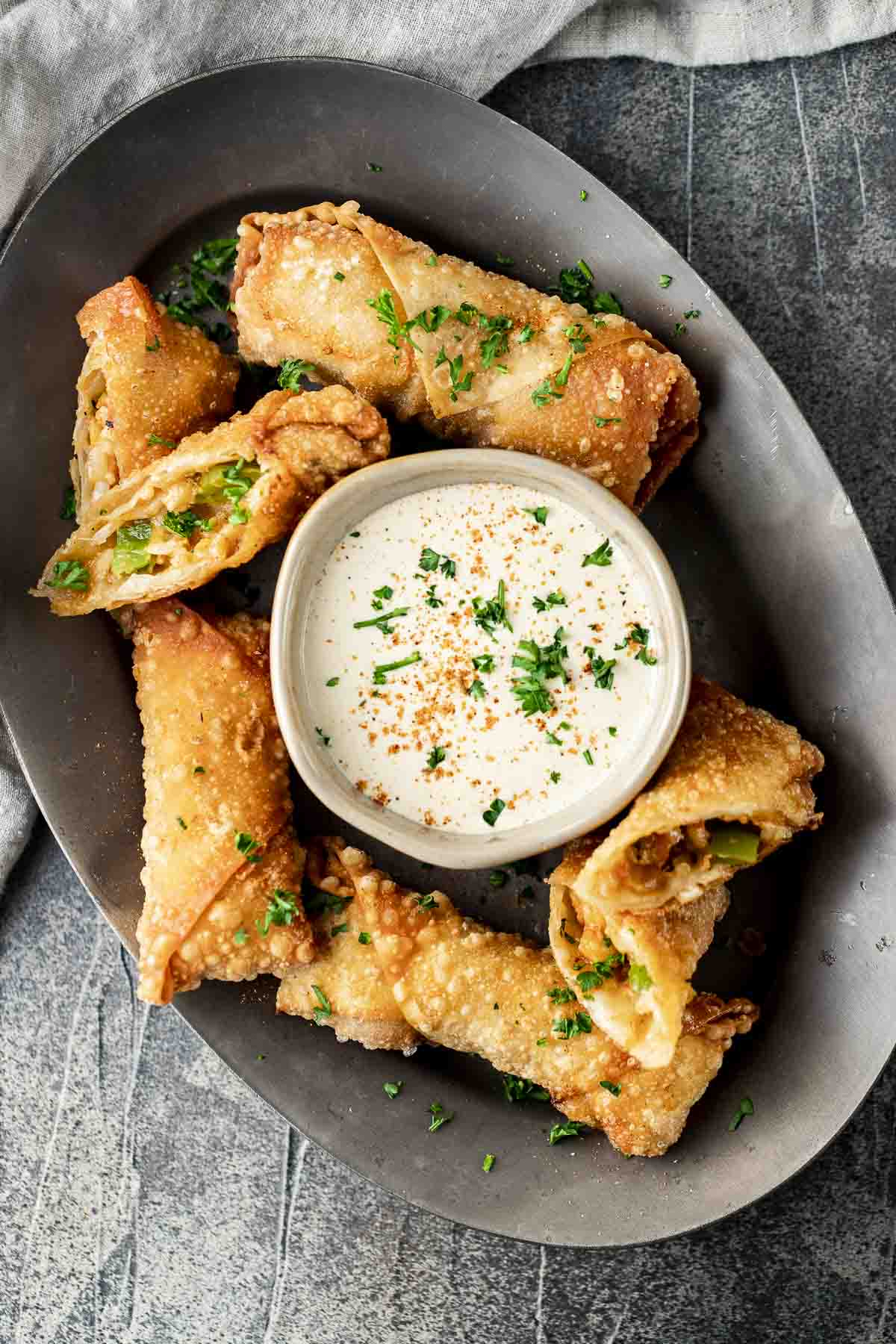a plate of egg rolls with white dipping sauce garnished with red pepper seasoning and parsley