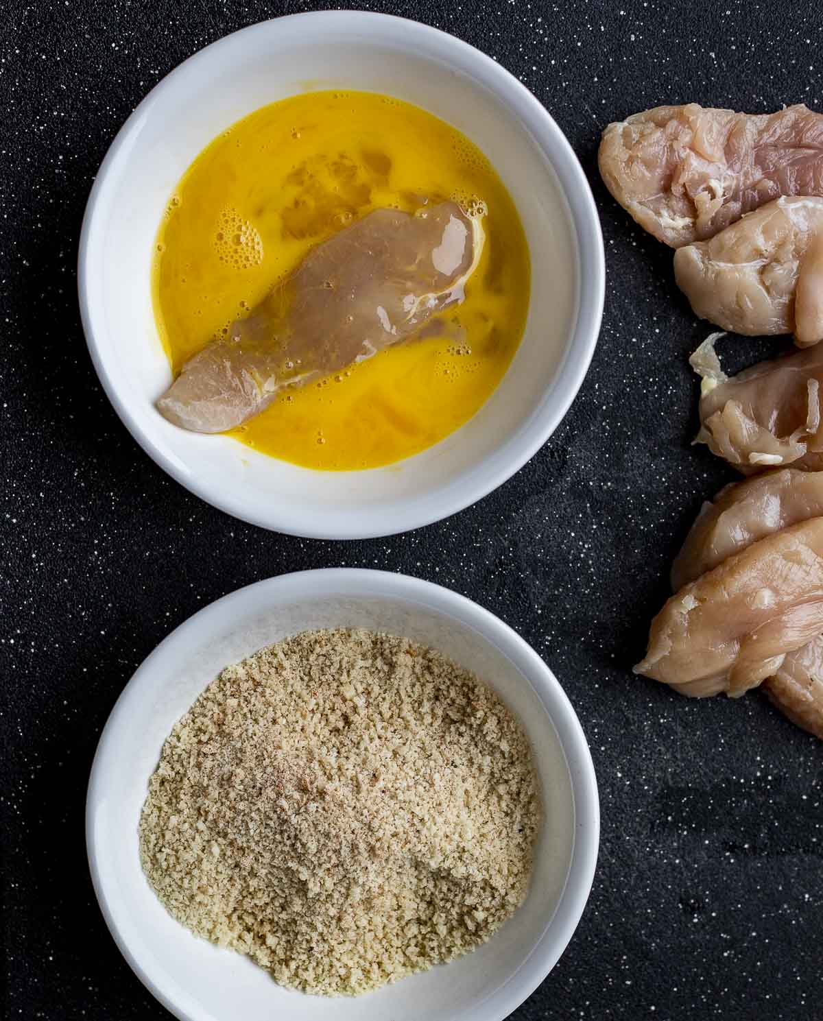 Chicken tenders being dipped in an egg wash next to a bowl of panko crumbs.