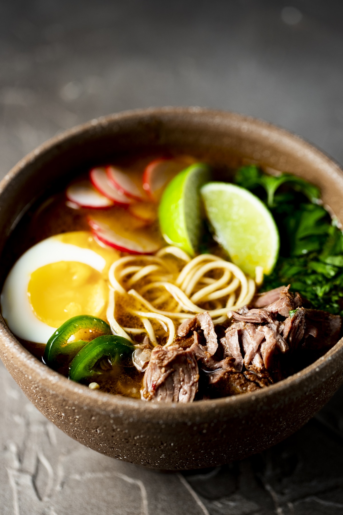 Birria ramen in a brown bowl with lime wedges and a ramen egg..