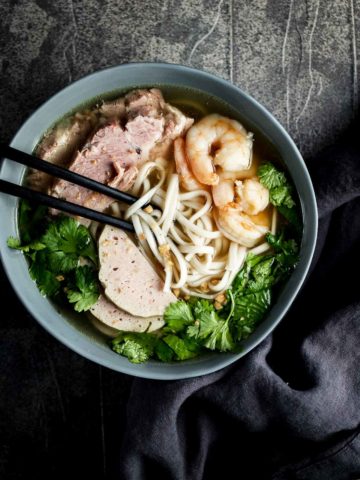 a bowl of noodles soup with chopsticks - sliced pork, shrimp and fresh green herbs