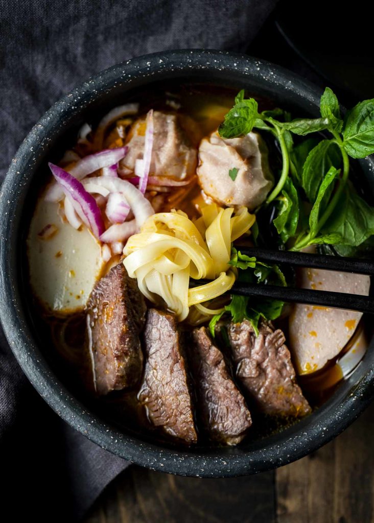 noodles being held by chopsticks in a bowl of beef soup with onions and fresh herbs