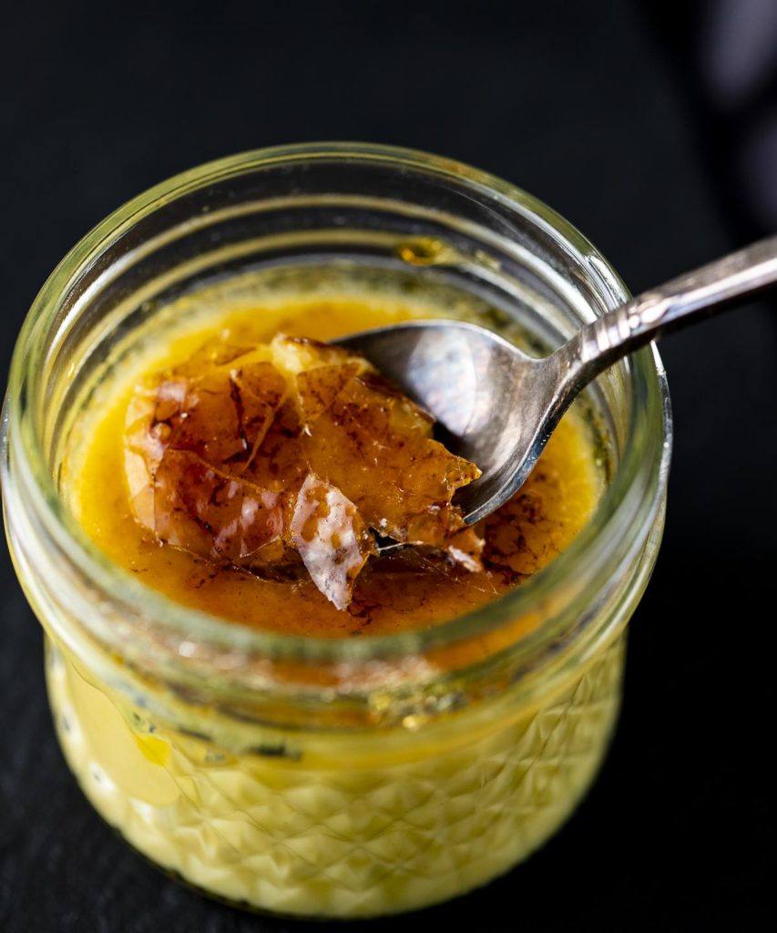 a spoon cracking the top of a glass jar of creme brulee