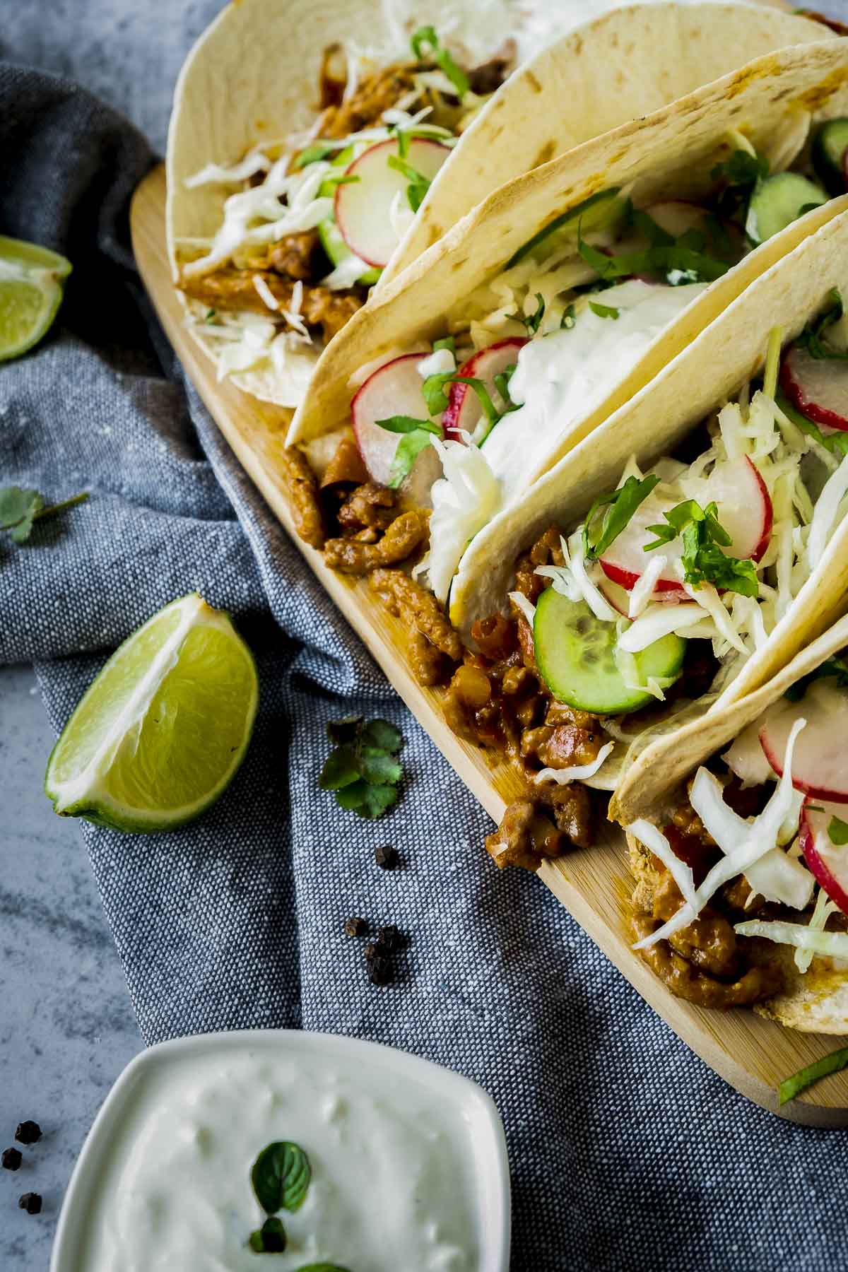 A serving board with four spicy lamb tacos beside a dipping sauce and lime.