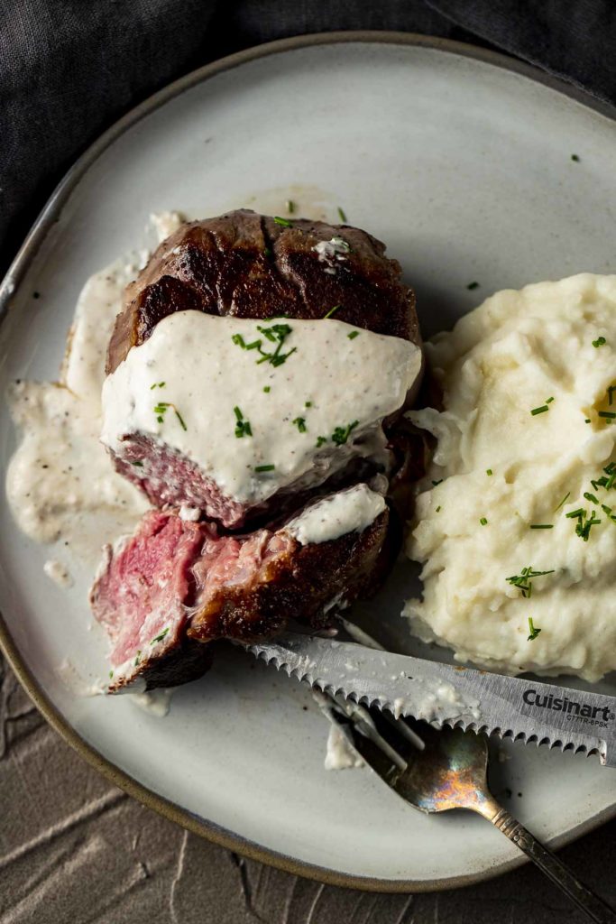 a piece of beef cut with a knife and covered in white sauce with mashed potatoes on the side
