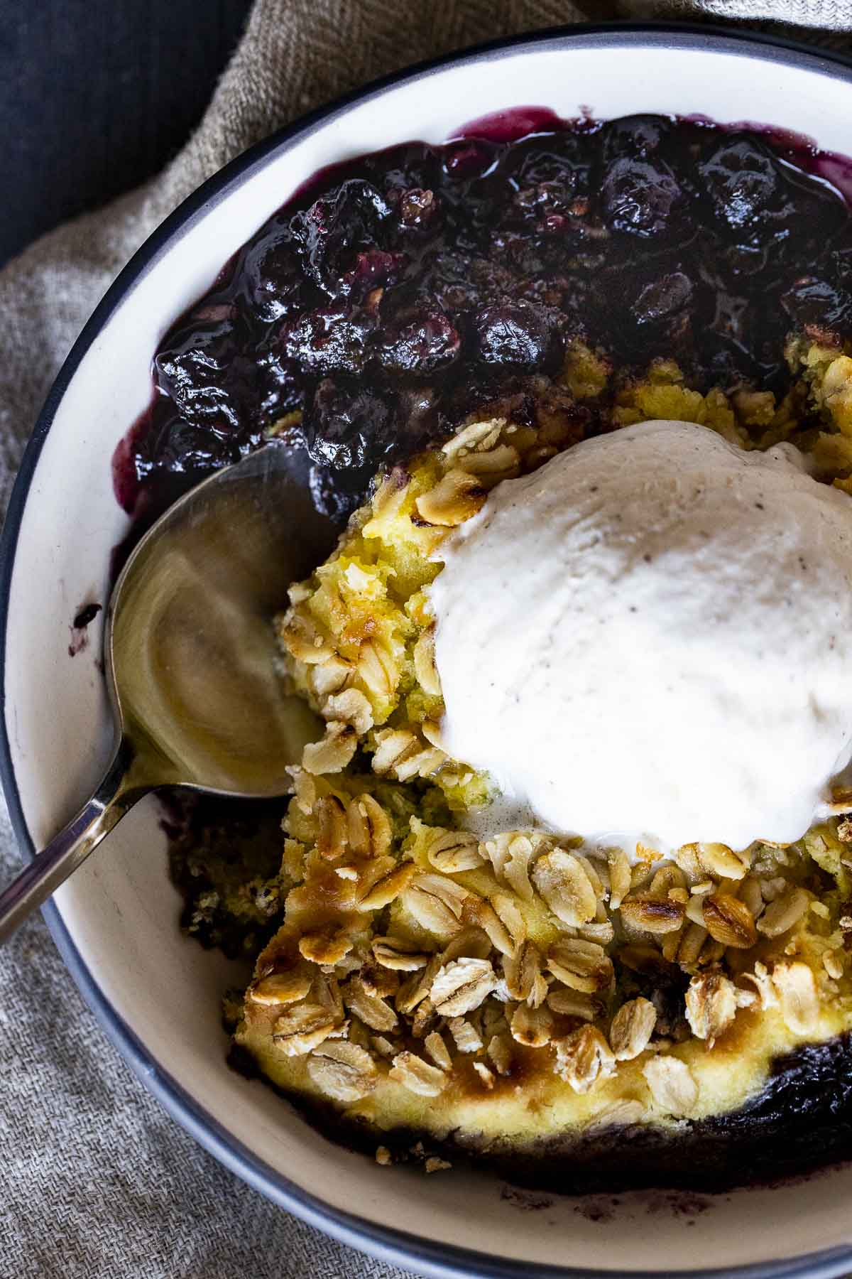 Overhead view of blueberry cobbler topped with ice cream and a large serving spoon inserted into it.