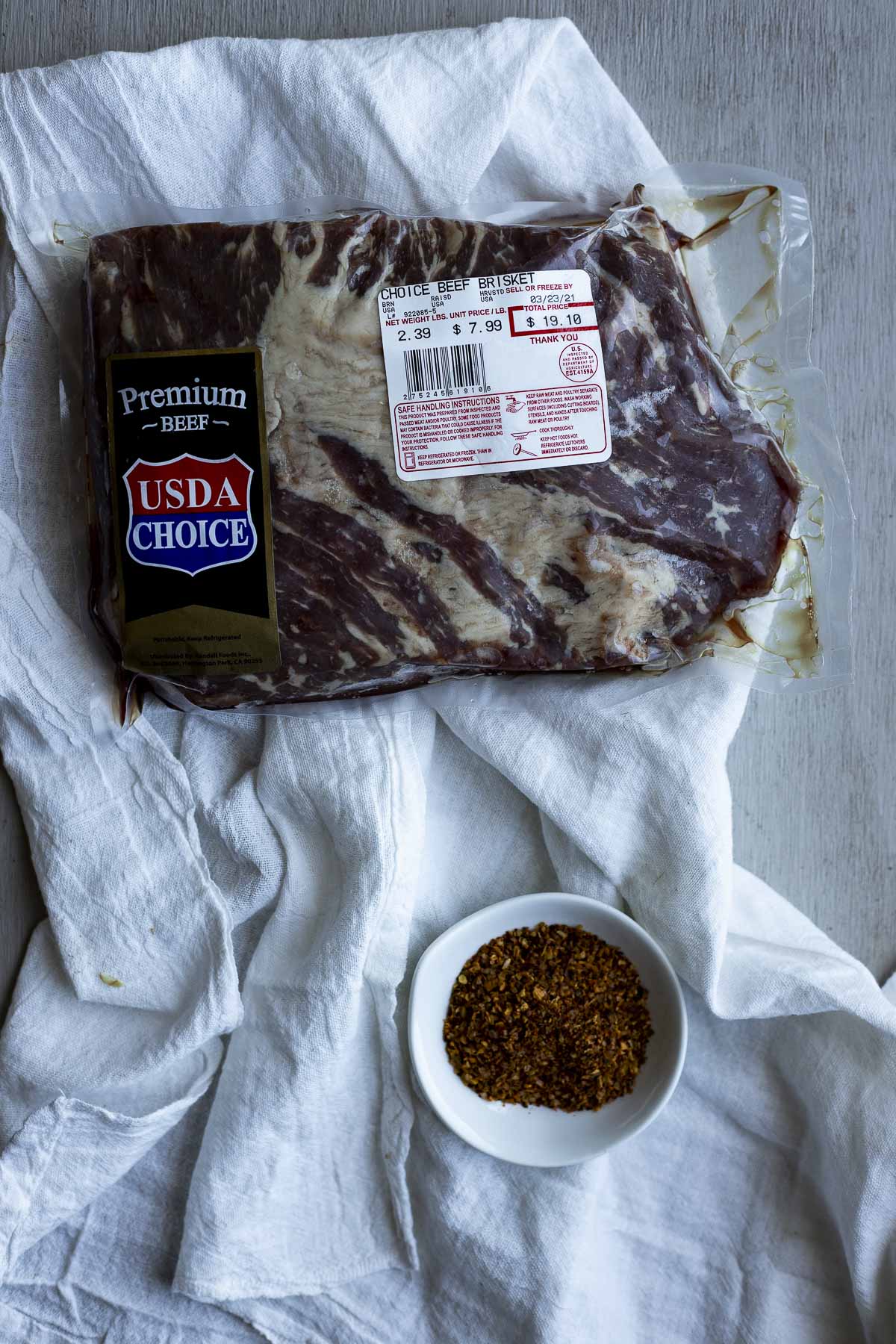 Brisket in packaging and a bowl of seasoning on a white cloth.