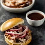 Pickled onions on top of pulled pork on a bun with BBQ sauce and pork in the background.