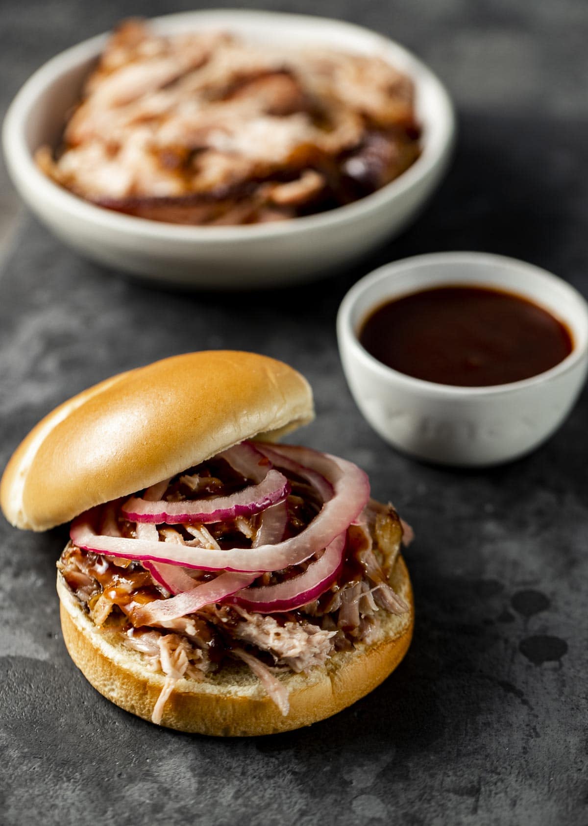 Pickled onions on top of pulled pork on a bun with BBQ sauce and pork in the background.