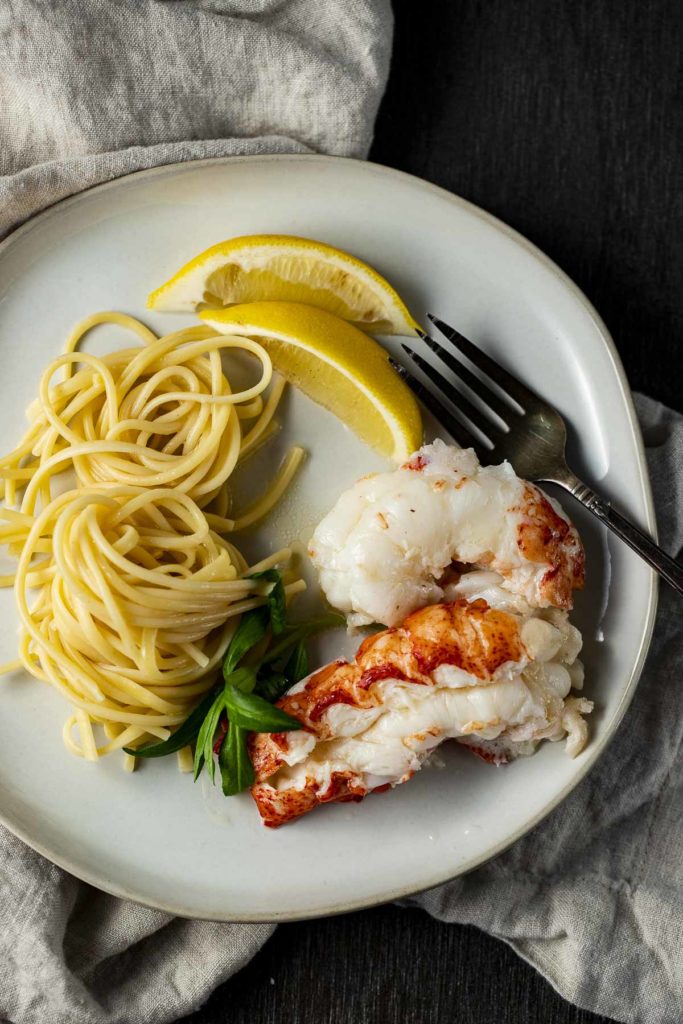 Overhead view of sous vide lobster tails, pasta and lemon wedges on a plate.