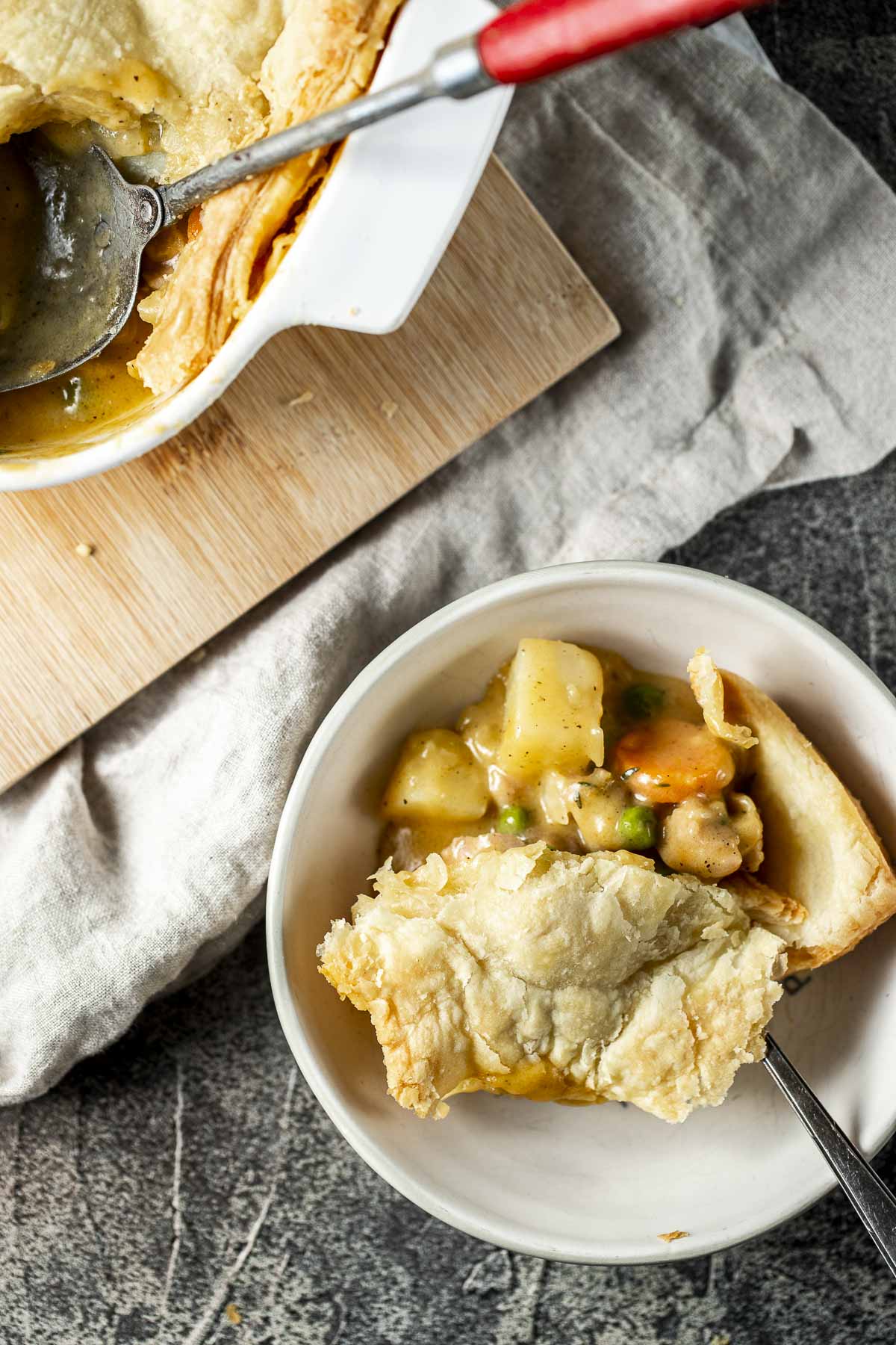 A piece of pot pie in a white bowl next to a larger dish of pot pie on a wooden board.