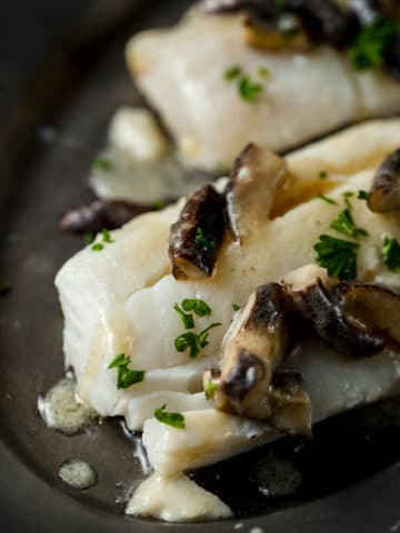 Close up view of a cod fillet with mushroom sauce and chopped herbs on top.