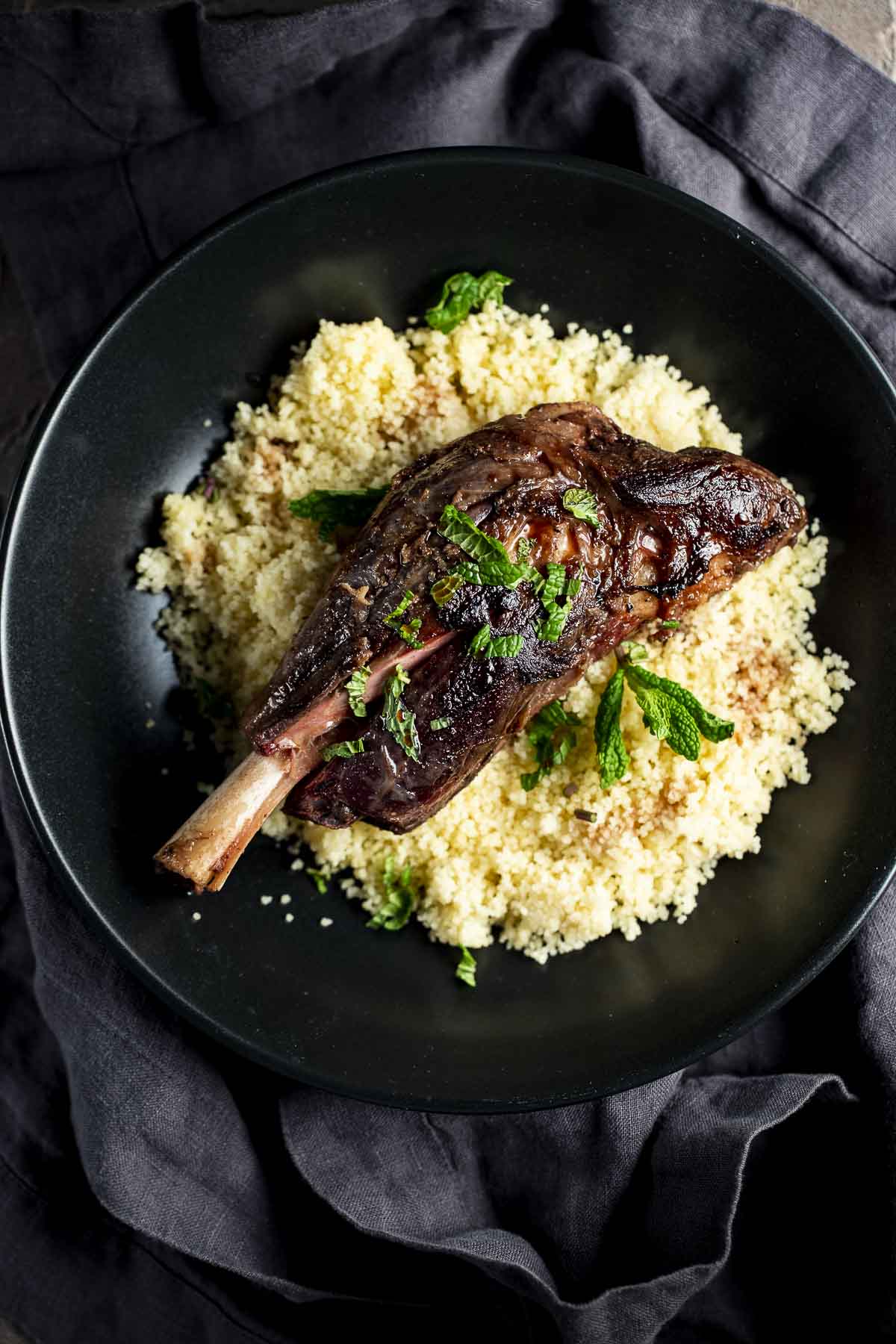 Overhead view of a lamb shank served on top of couscous on a black plate.