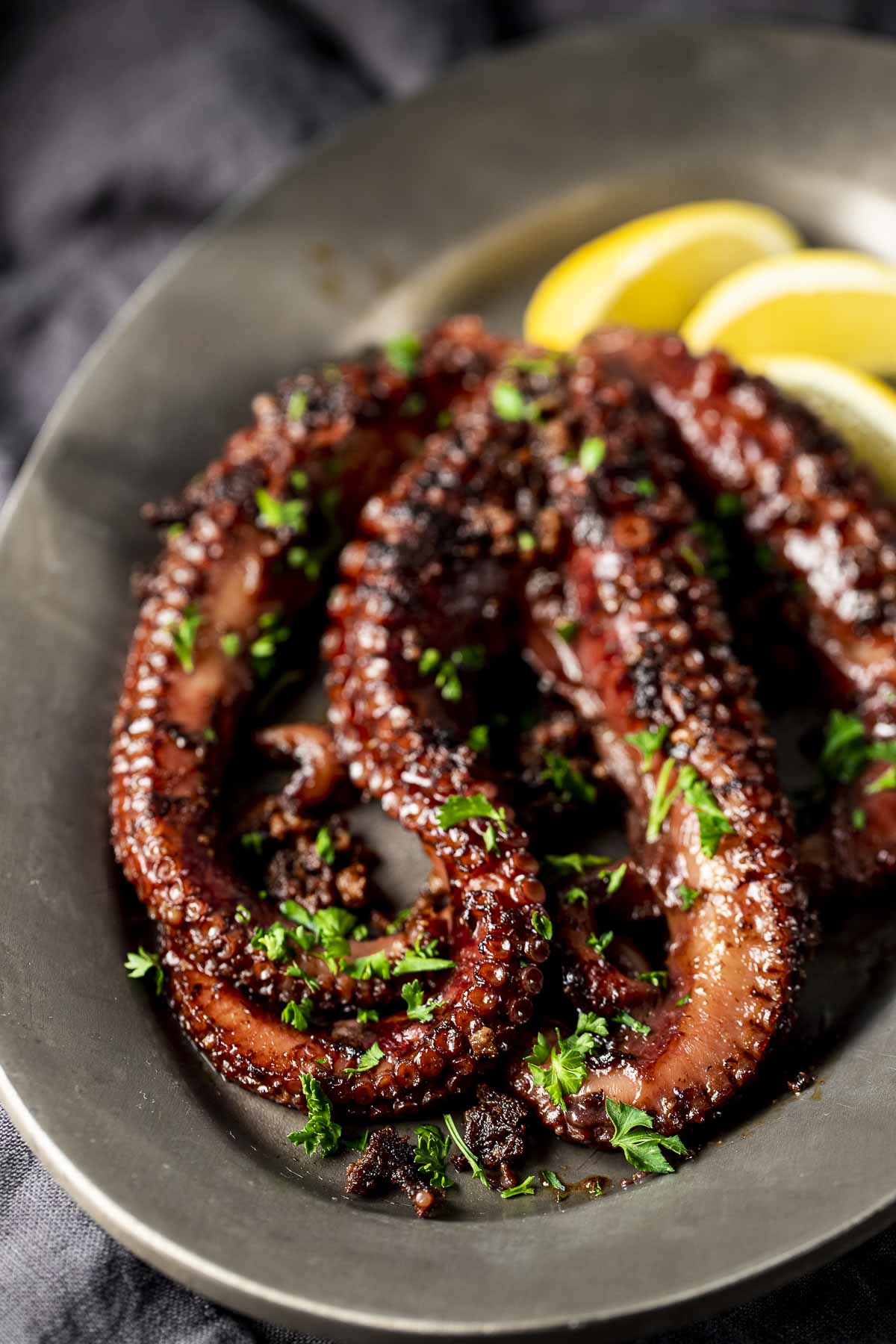 Octopus topped with chopped herbs on a grey plate with lemon wedges.