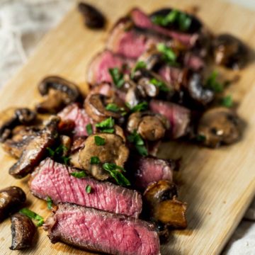 Sirloin steak cut into slices on a wooden board and topped with mushroom sauce.