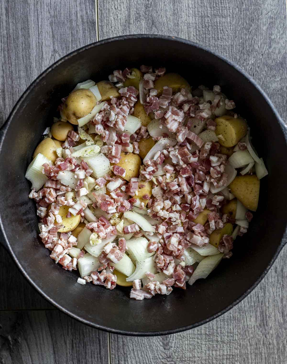 Mini potatoes, onions, garlic and pancetta arranged in the bottom of a Dutch oven.