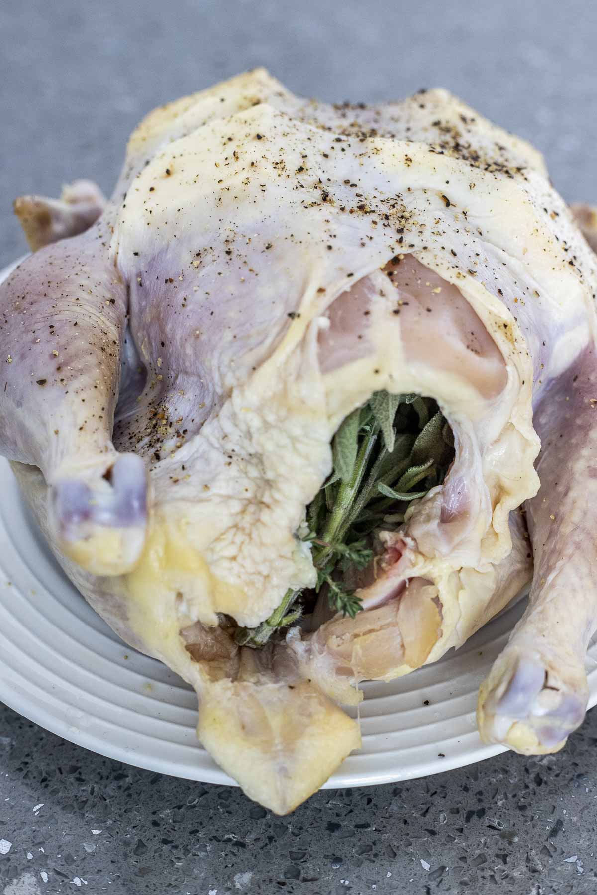 Whole raw chicken on a white plate with butter pieces under the skin and fresh herbs visible in the cavity.