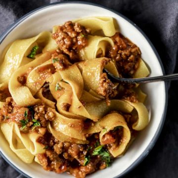 Bolognese sauce mixed in with pappardelle pasta in a white bowl on a grey cloth.