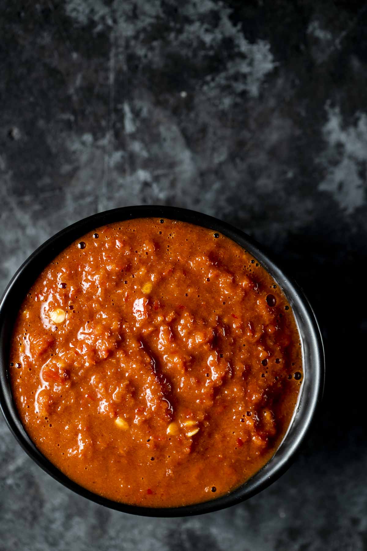 Overhead view of sambal belacan in a black bowl on a dark surface.