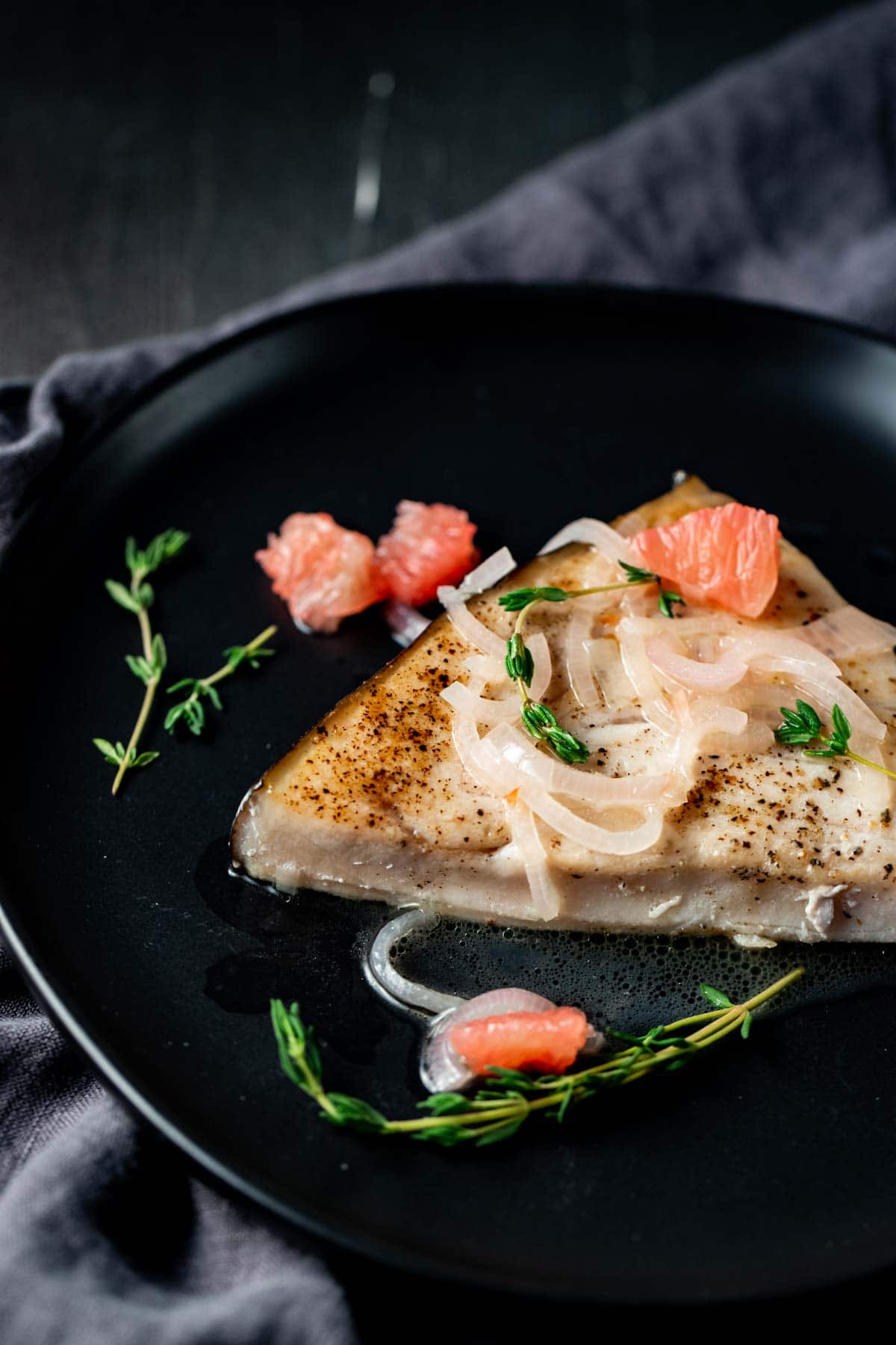 A piece of sous vide swordfish on a black plate with grapefruit and shallot sauce on top.