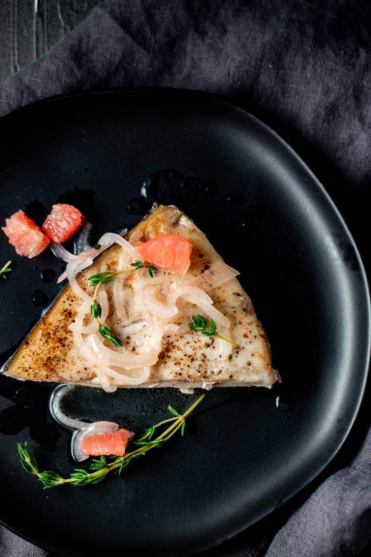 Overhead view of a swordfish steak on a black plate with shallots and grapefruit pieces on top.