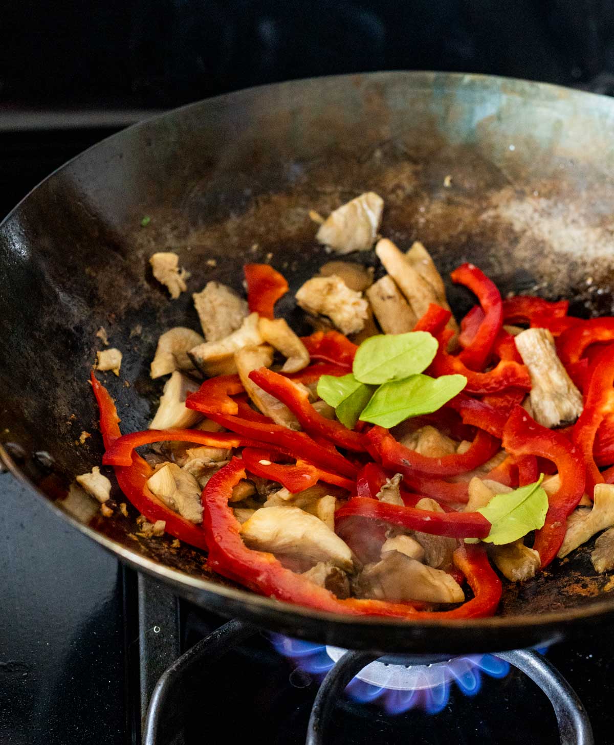 Red peppers, mushrooms and lime leaves cooking in a wok on the stovetop.