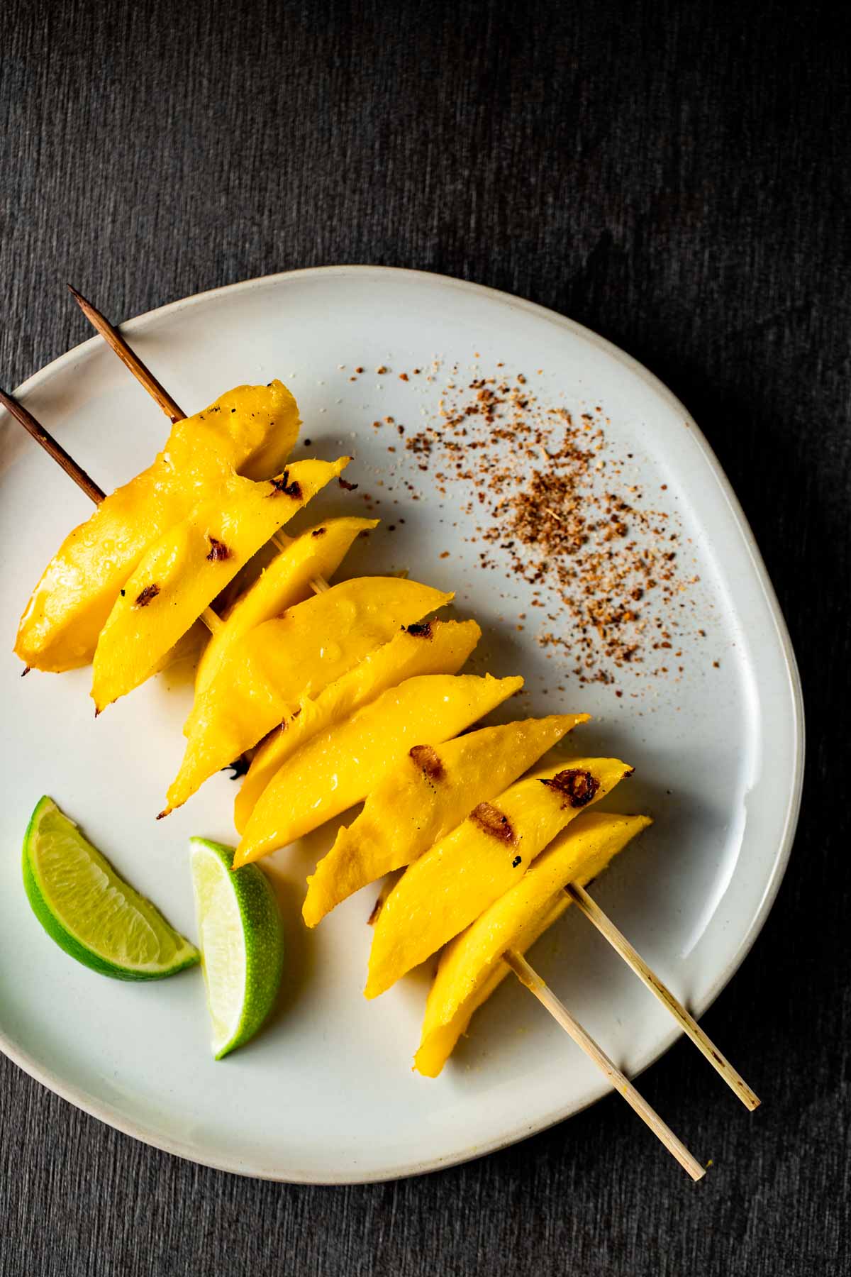 Grilled mango slices on skewers on a white plate with lime slices and tajin on the side.