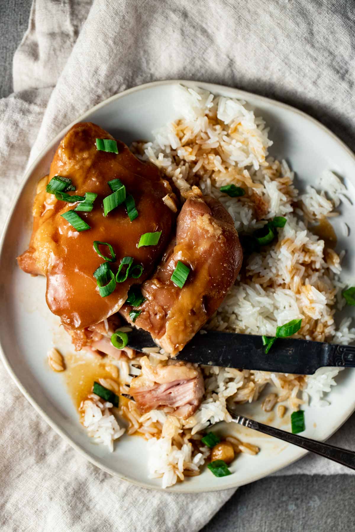 A teriyaki chicken thigh being cut with a fork and knife on a plate.