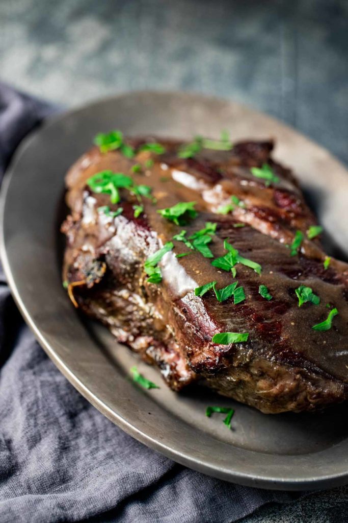 Side view of a pot roast on a platter and topped with gravy and chopped herbs.