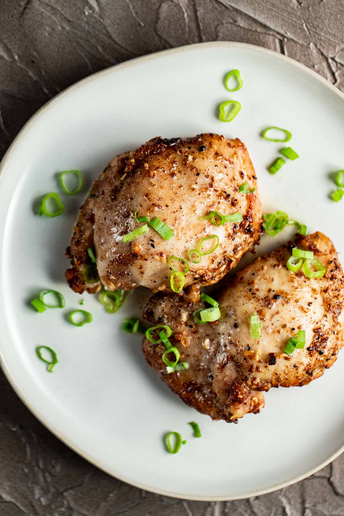 Overhead view of boneless chicken thighs on a white plate with chopped green onions on top.