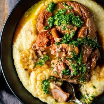 Overhead view of osso buco and gremolata served on polenta in a black bowl with a spoon inserted into it.