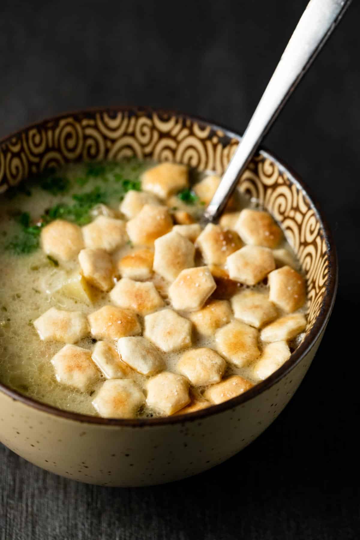 Clam chowder in a bowl with a spoon inserted into it and oyster crackers on top.