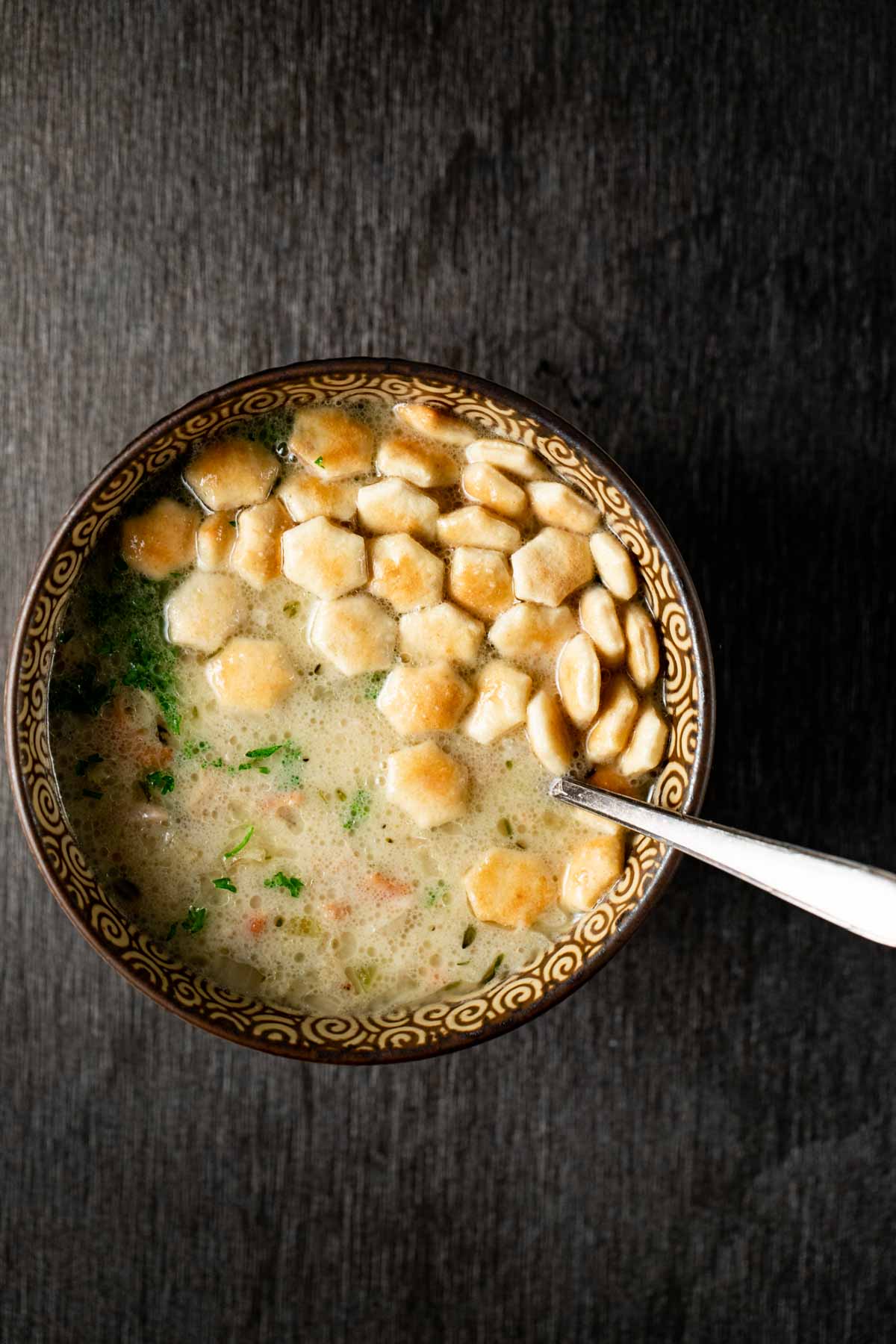 Overhead view of a bowl of clam chowder with crackers on top and a spoon inserted into the bowl.