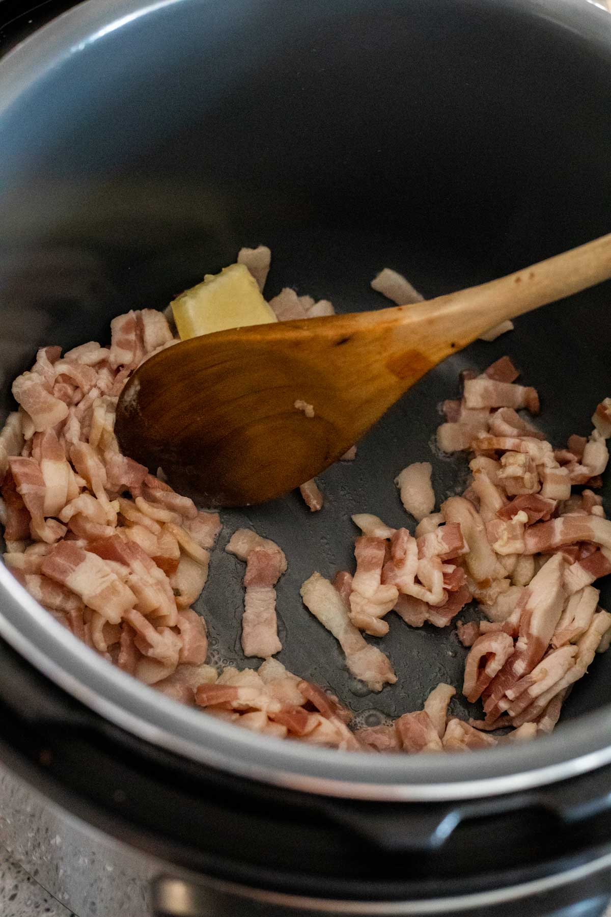 Bacon with butter being stirred around in the Instant Pot insert with a wooden spoon.