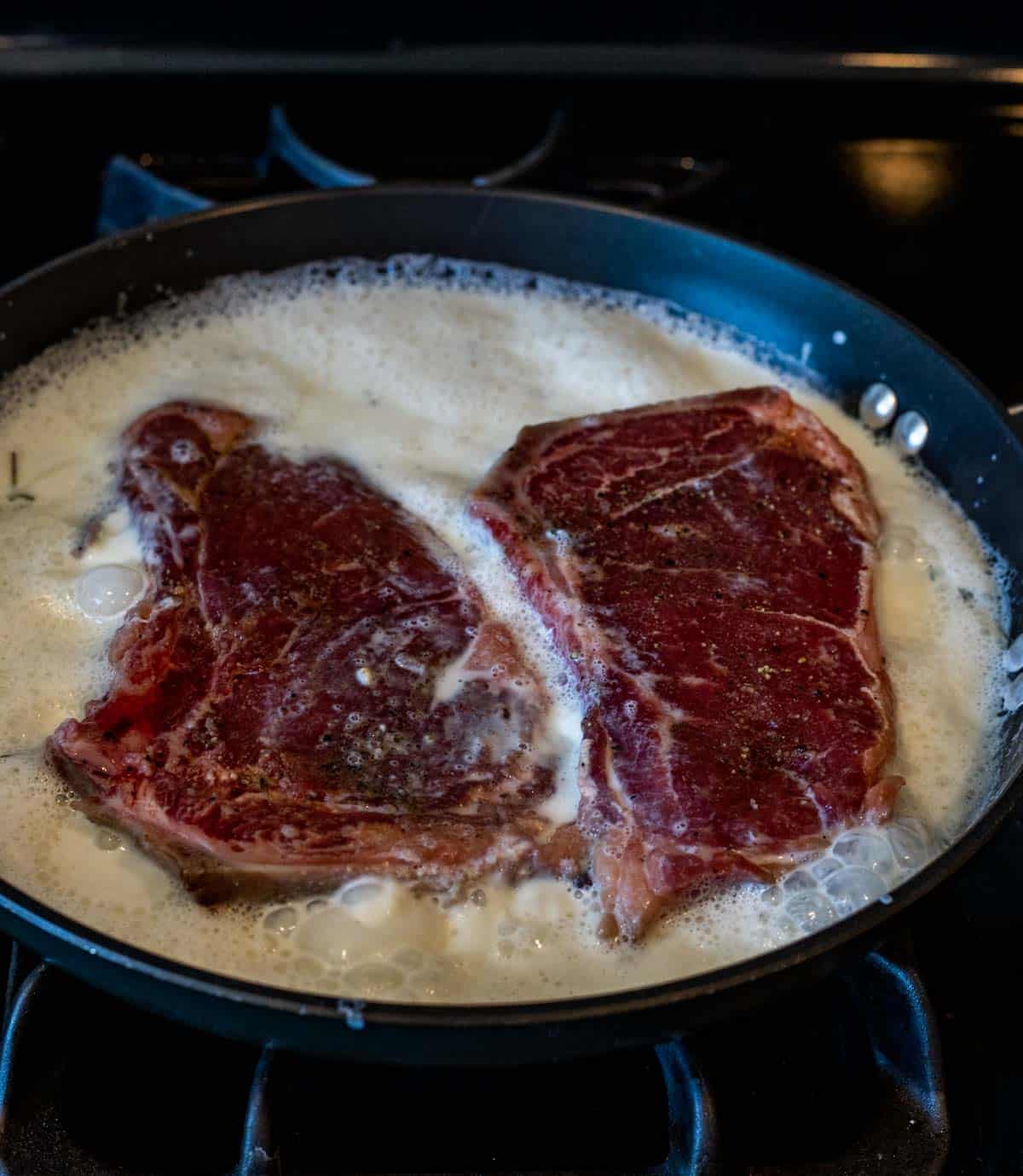 Steaks boiling in milk in a skillet on the stovetop.