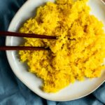 Overhead view of turmeric rice on a white plate with chopsticks and resting on a blue cloth.