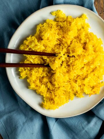 Overhead view of turmeric rice on a white plate with chopsticks and resting on a blue cloth.