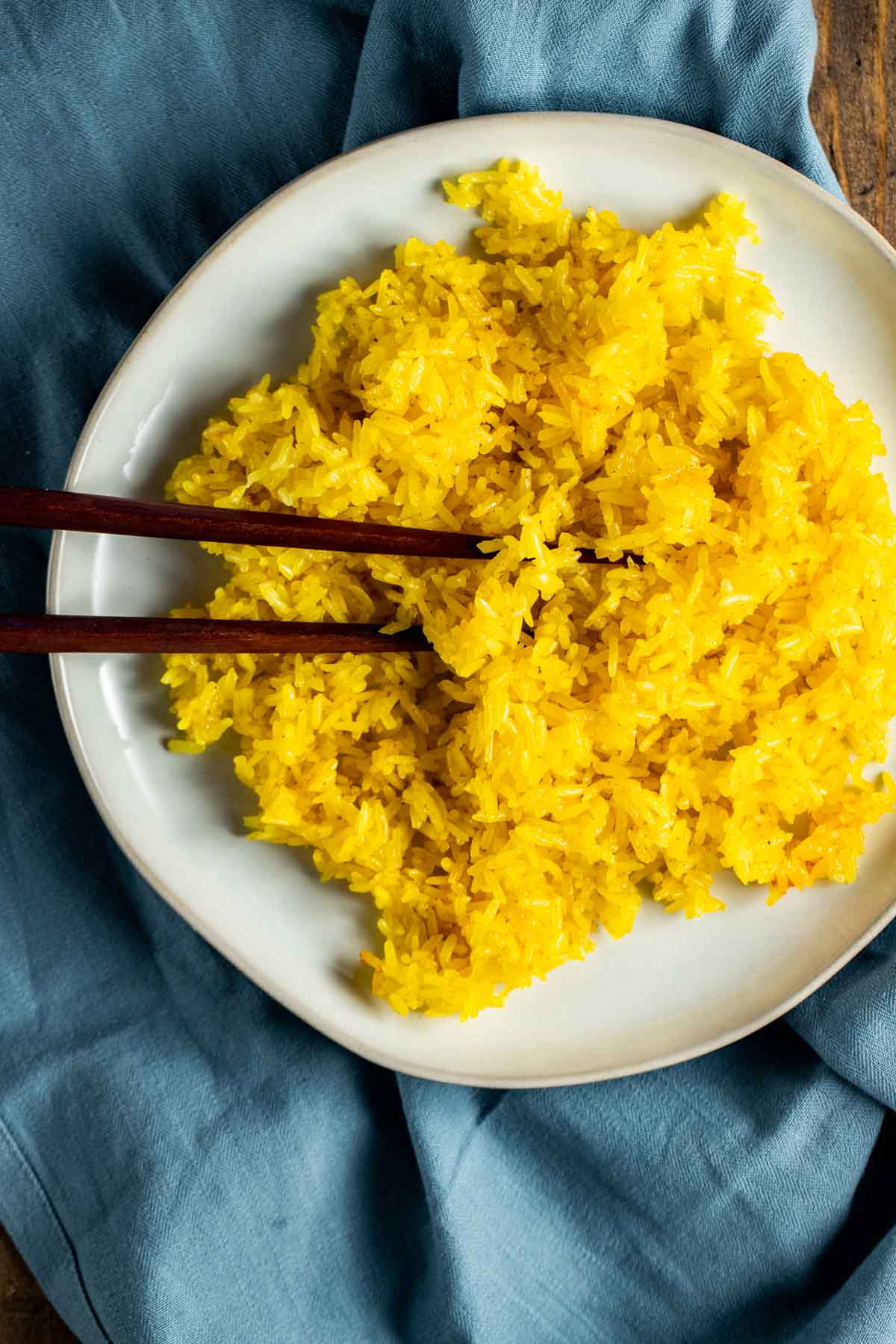 Overhead view of turmeric rice on a white plate with chopsticks and resting on a blue cloth.