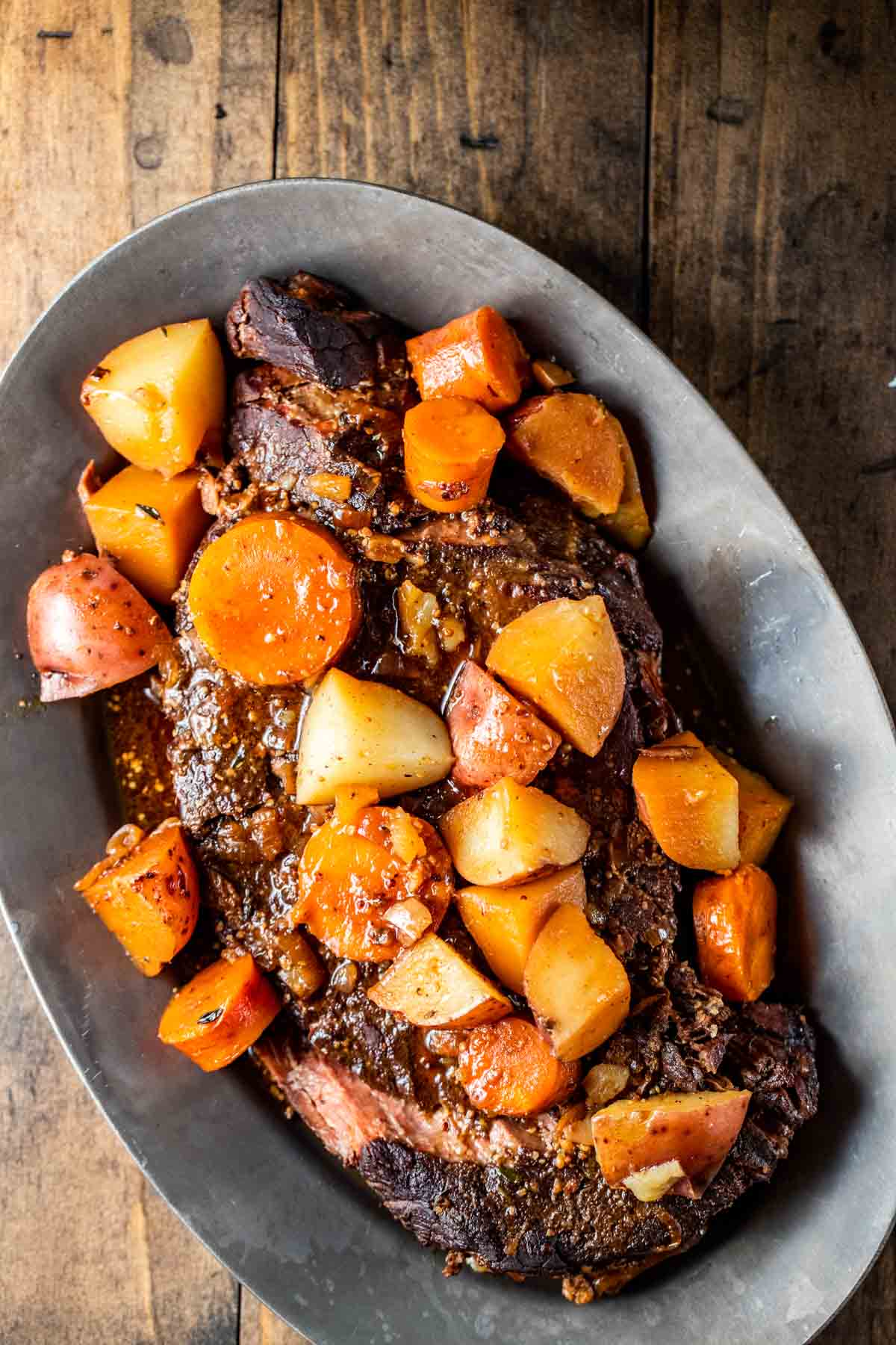 a large cooked piece of beef on a silver platter served with cooked carrots and potatoes