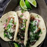 Three tortillas on a platter and filled with carne asada and toppings with lime wedges on the side.