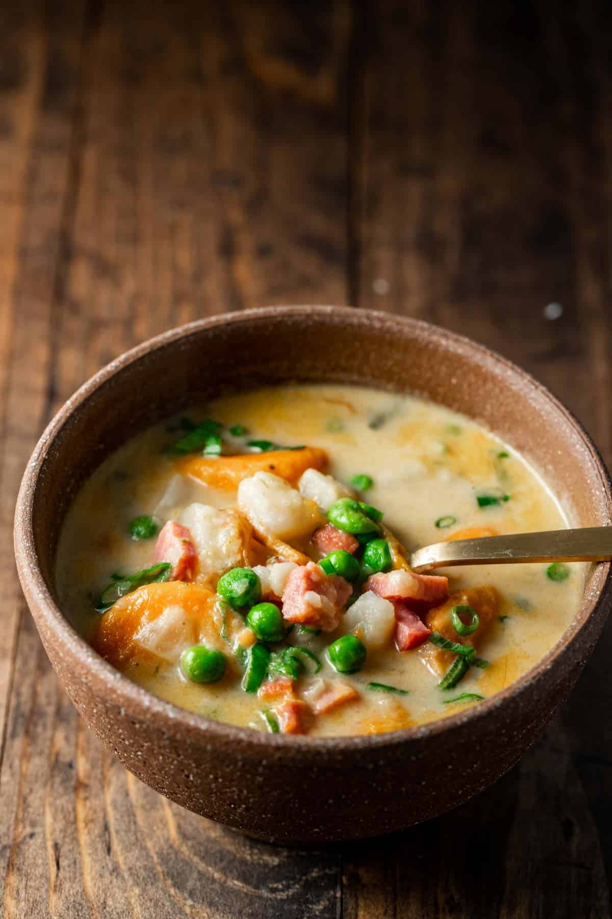 Ham and potato soup in a brown bowl with a spoon and resting on a brown wooden surface.