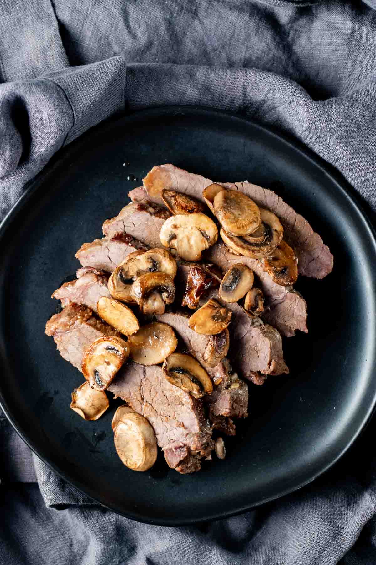 Overhead view of sliced London broil topped with mushrooms on a black plate.