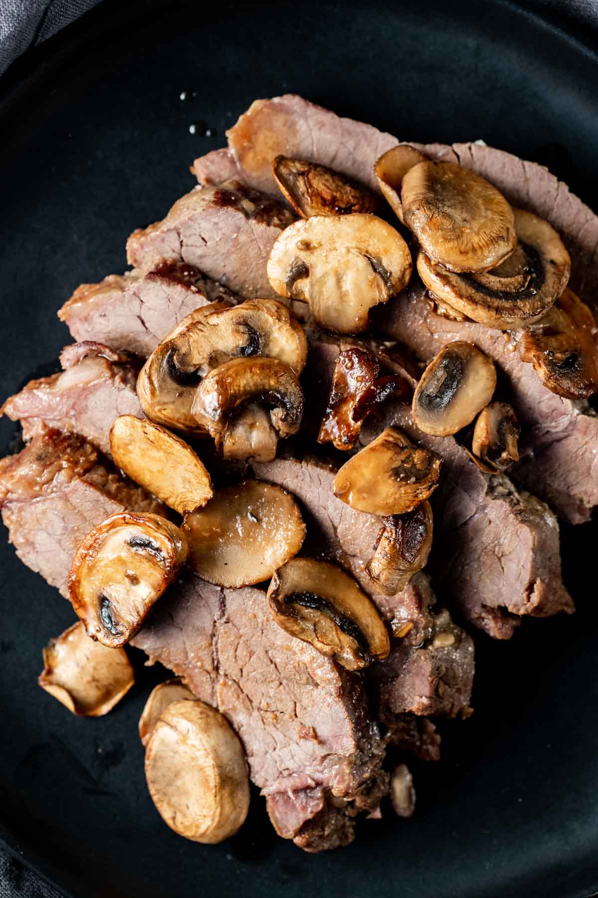 London broil topped with mushrooms and served on a black plate.