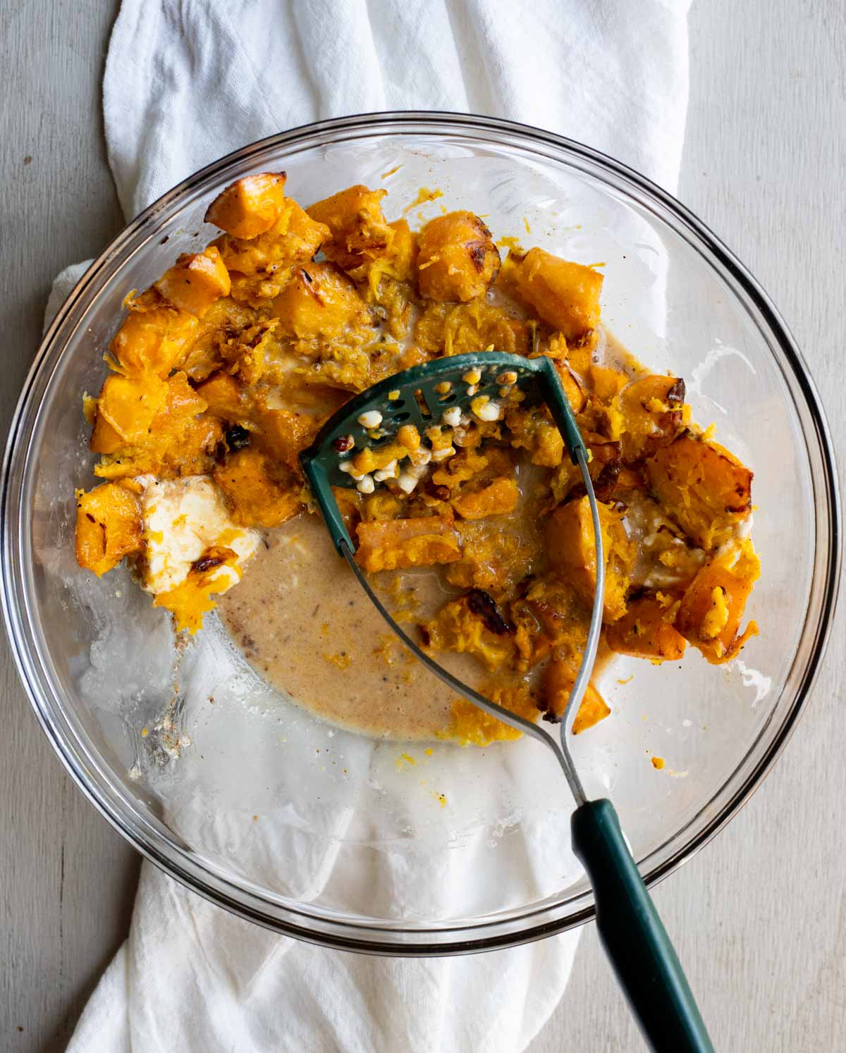Roasted pumpkin cubes in a glass bowl and being mashed with a potato mashed.